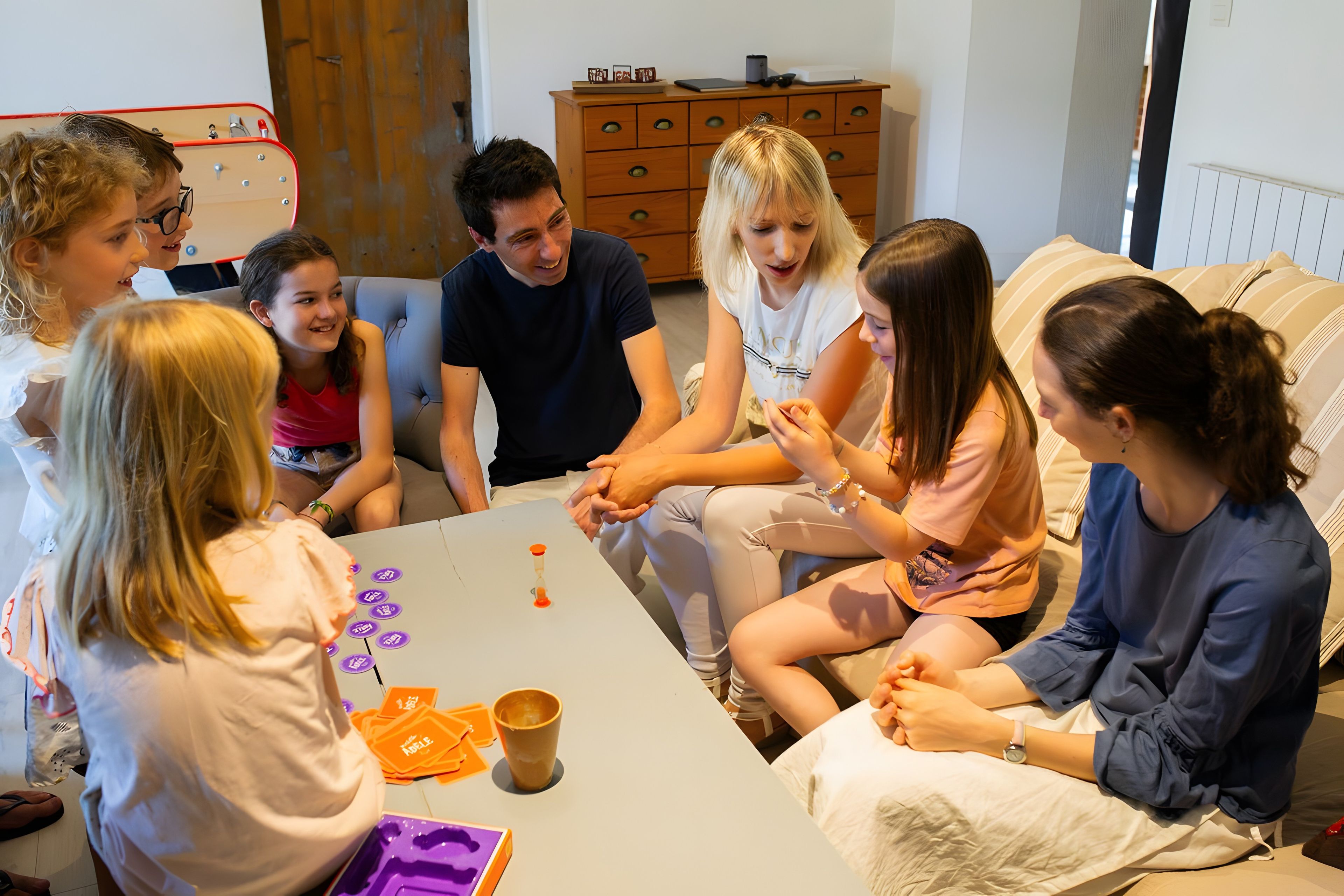 A group of people seated around a table with cards and a cup, in a room with a couch and a wooden cabinet.