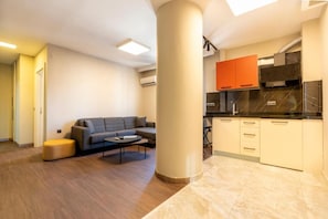A modern living room with a sofa and ottoman, a kitchen with black countertops and orange cabinets, and a large pillar. - Premium Rezidans Otel (Muratpasa)