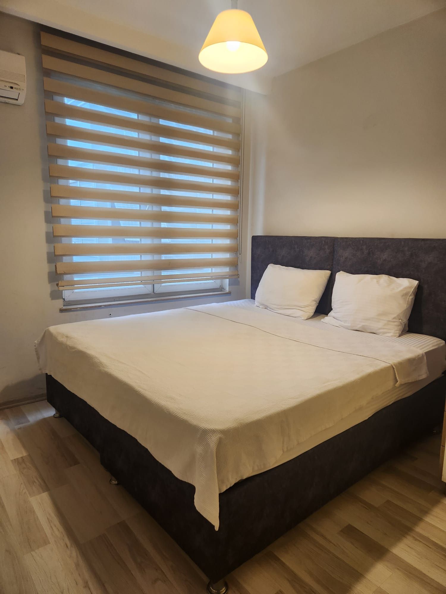 A bedroom with a bed, a window with blinds, a ceiling light, and a wall-mounted air conditioning unit.