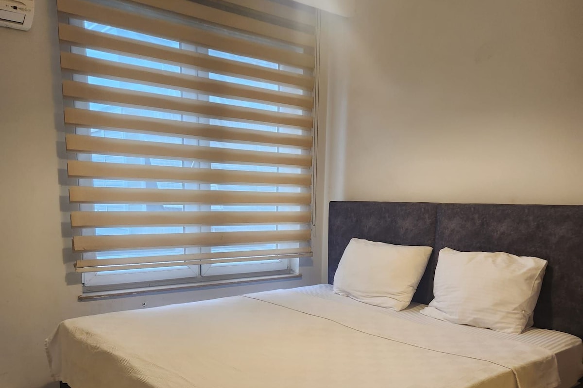 A bedroom with a bed, a window with blinds, a ceiling light, and a wall-mounted air conditioning unit.