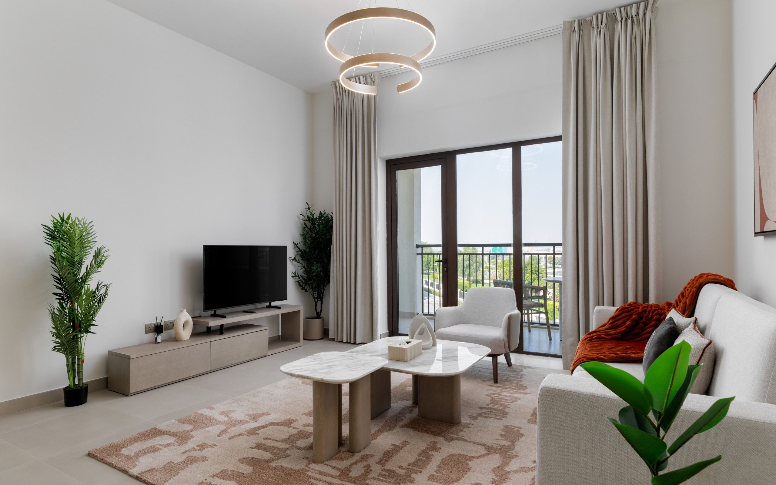 A modern living room with a flat-screen TV, a sofa, a coffee table, and a balcony with curtains.