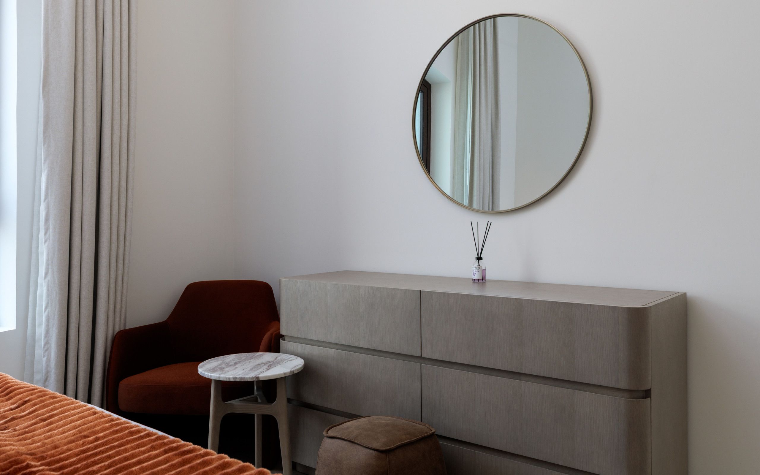 A hotel room with a single bed, a small round table, a red chair, a grey cabinet, and a round mirror on the wall.