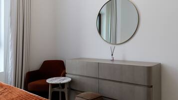 A hotel room with a single bed, a small round table, a red chair, a grey cabinet, and a round mirror on the wall.