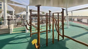 A children's play area with a climbing structure and a slide, surrounded by outdoor seating and a view of the city skyline.