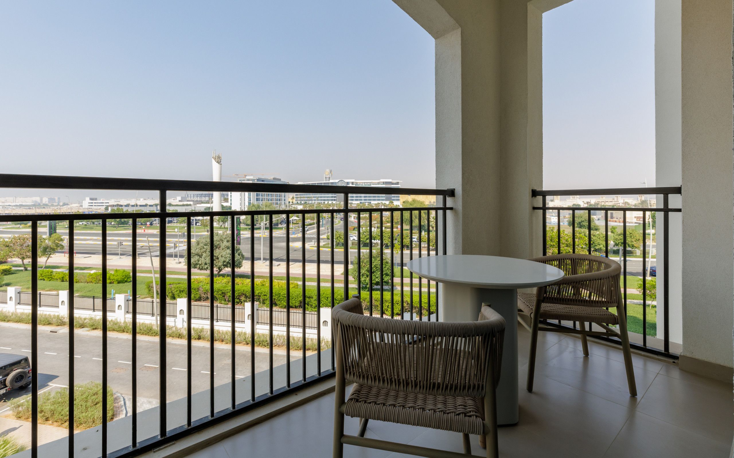 A balcony with a table and two wicker chairs, overlooking a road and buildings.