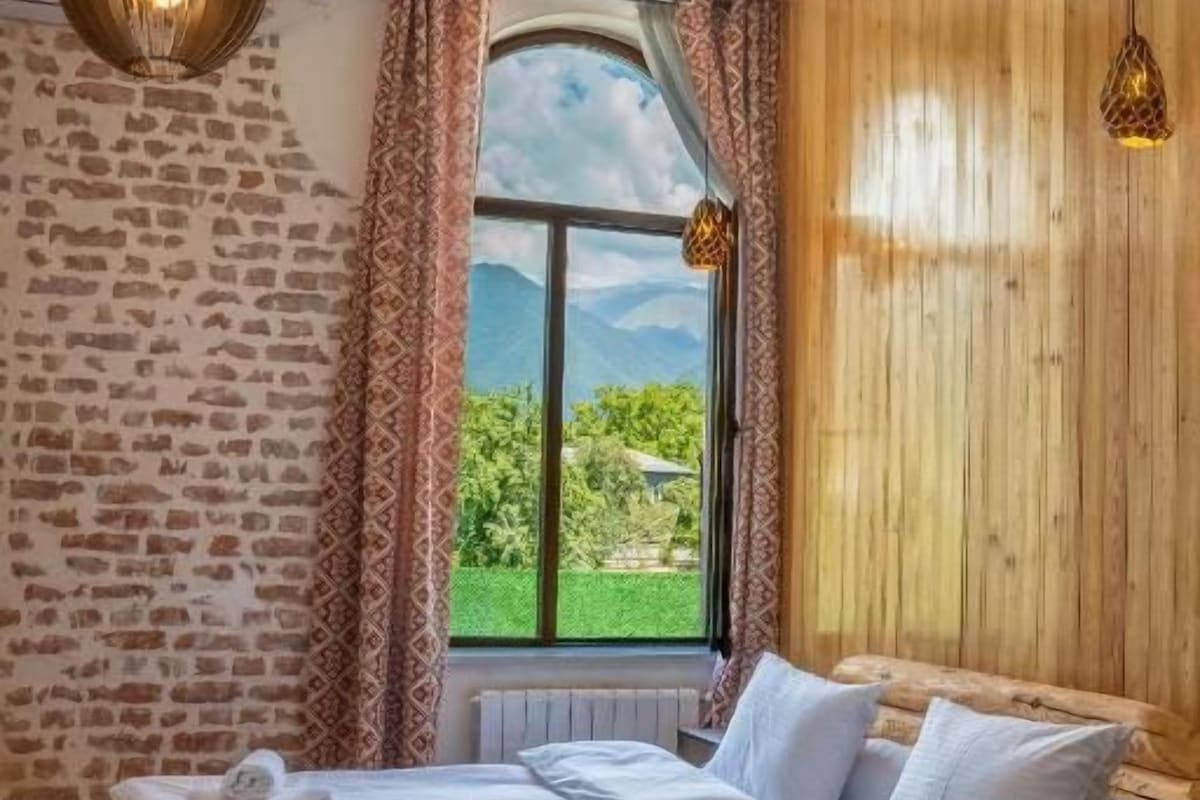 A room with a wooden bed, a window with curtains, and a view of greenery.