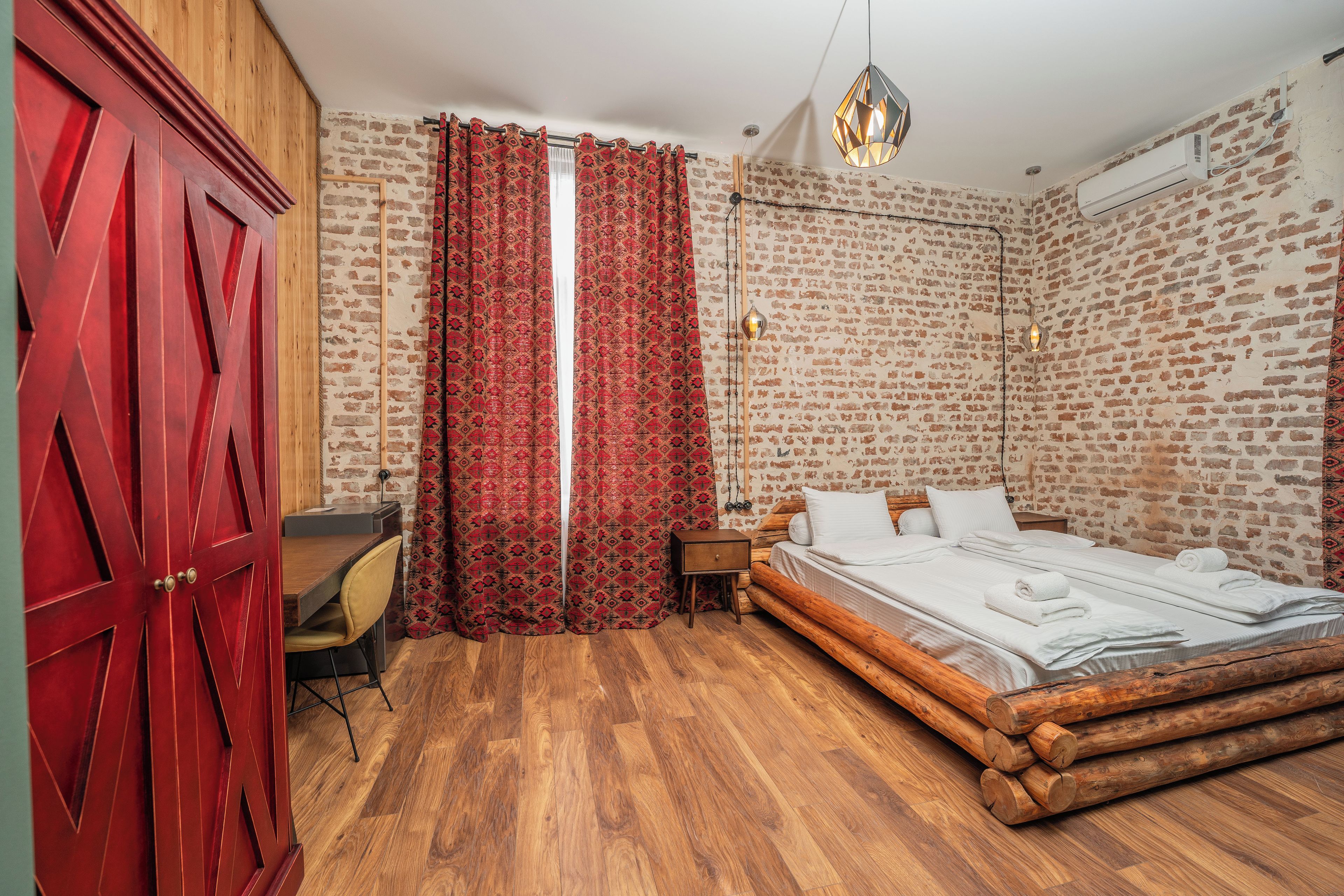 A room with a wooden floor, a bed with white bedding, a desk with a chair, a wardrobe, and a brick-patterned wall.