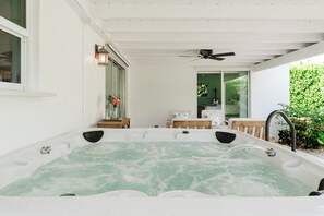 A hot tub with a wooden deck and a ceiling fan, surrounded by a white exterior wall and a glass door leading to a green garden. - Juno Breeze | Hot Tub | Walk to Beach | Sleeps 9! (Juno Beach)