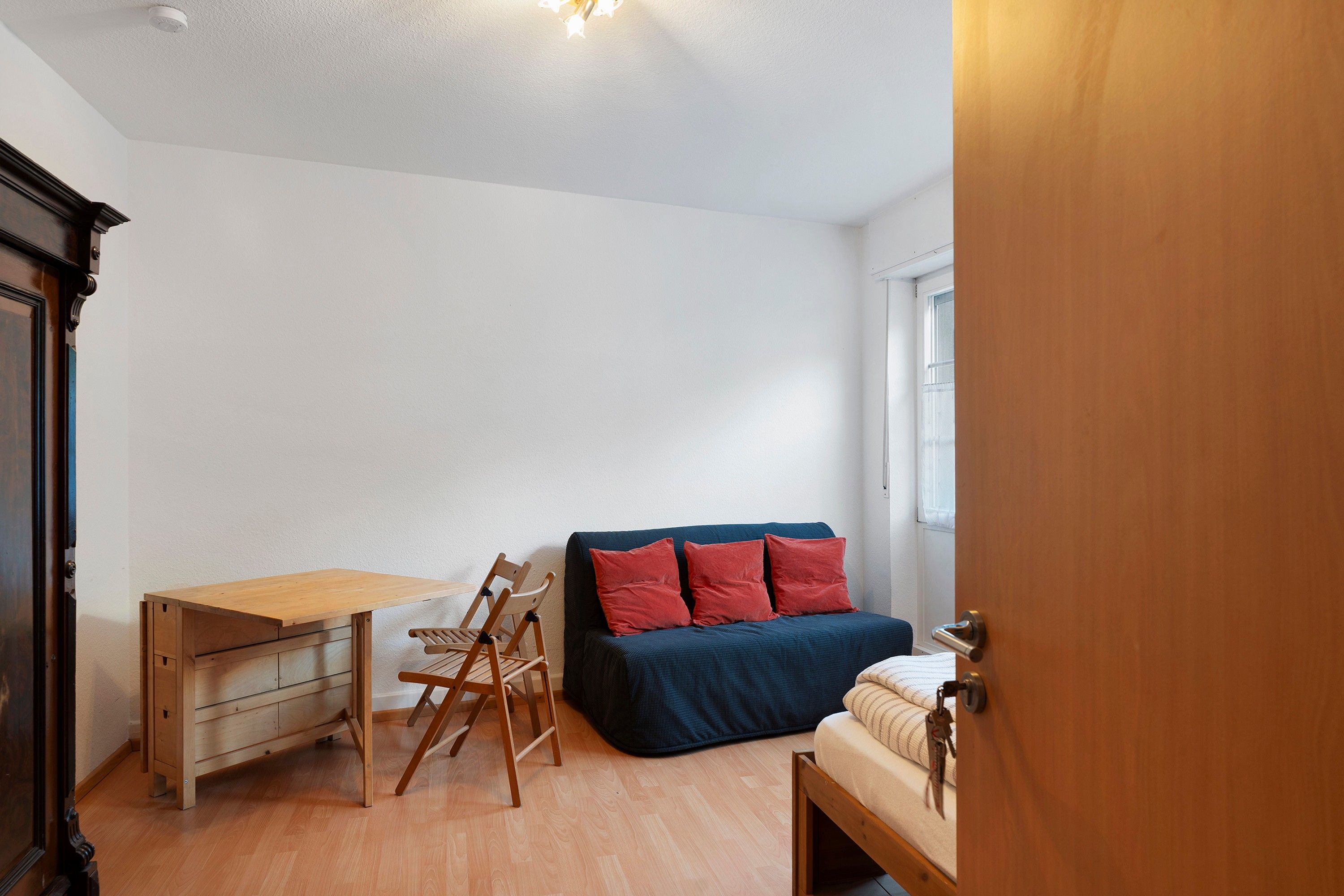 Una habitación con un escritorio de madera, un sofá azul con cojines rojos y una mesita de noche pequeña.