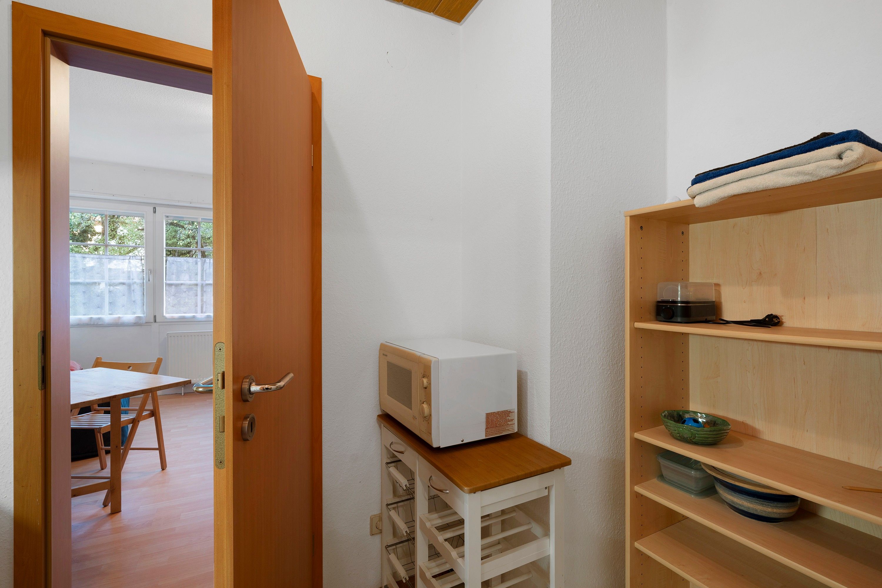 A room with a wooden door, a white wall, a microwave on a small wooden shelf, and a window with a view of greenery outside.
