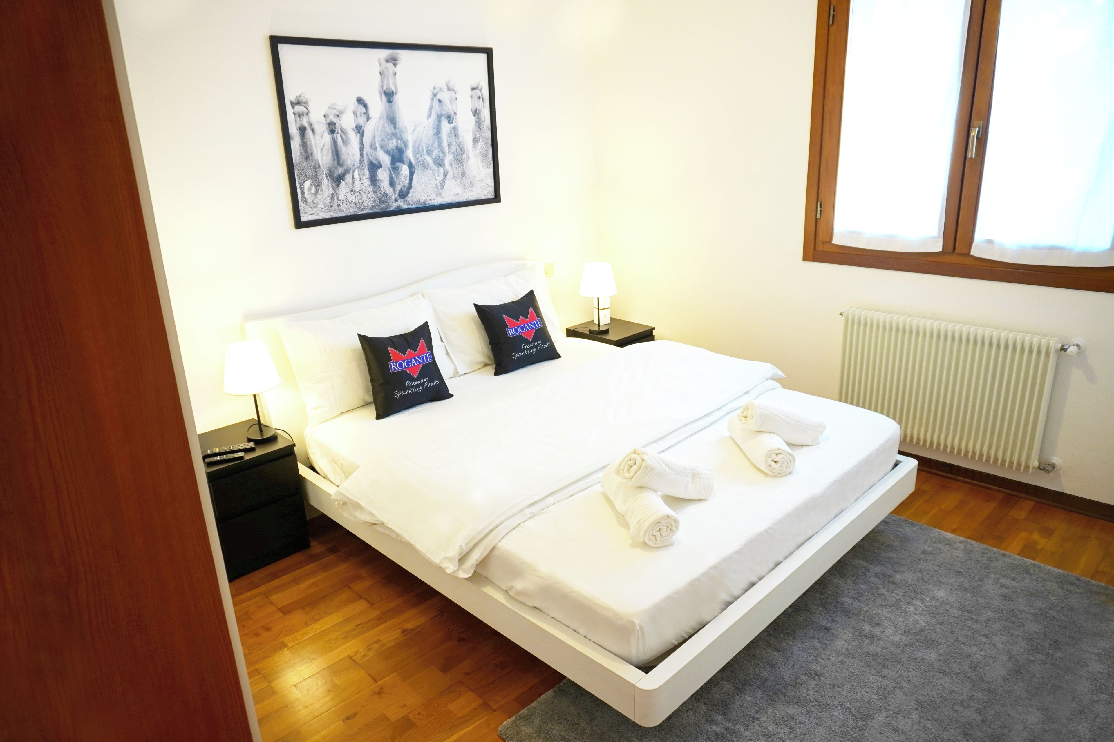 Una camera d'albergo con un letto, cuscini, un comodino, una lampada, un quadro appeso al muro e una finestra.