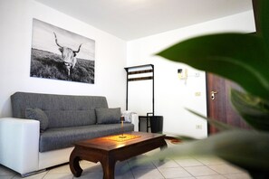 A living room with a grey sofa, a wooden coffee table, a black floor lamp, and a wall-mounted light fixture. - Comfortable Apartment, Near CRO and the U.S. Base (Castello d'Aviano)