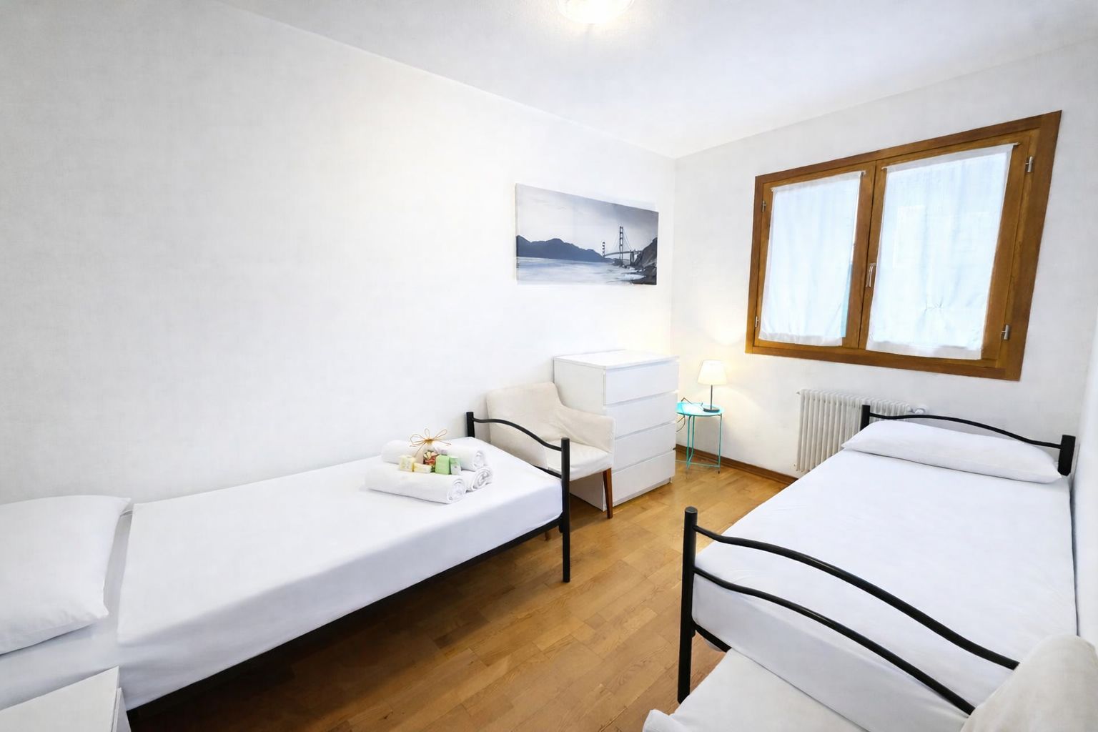 A room with two single beds, a small desk, a chair, a radiator, and a window with curtains.