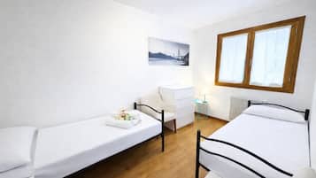A room with two single beds, a small desk, a chair, a radiator, and a window with curtains.