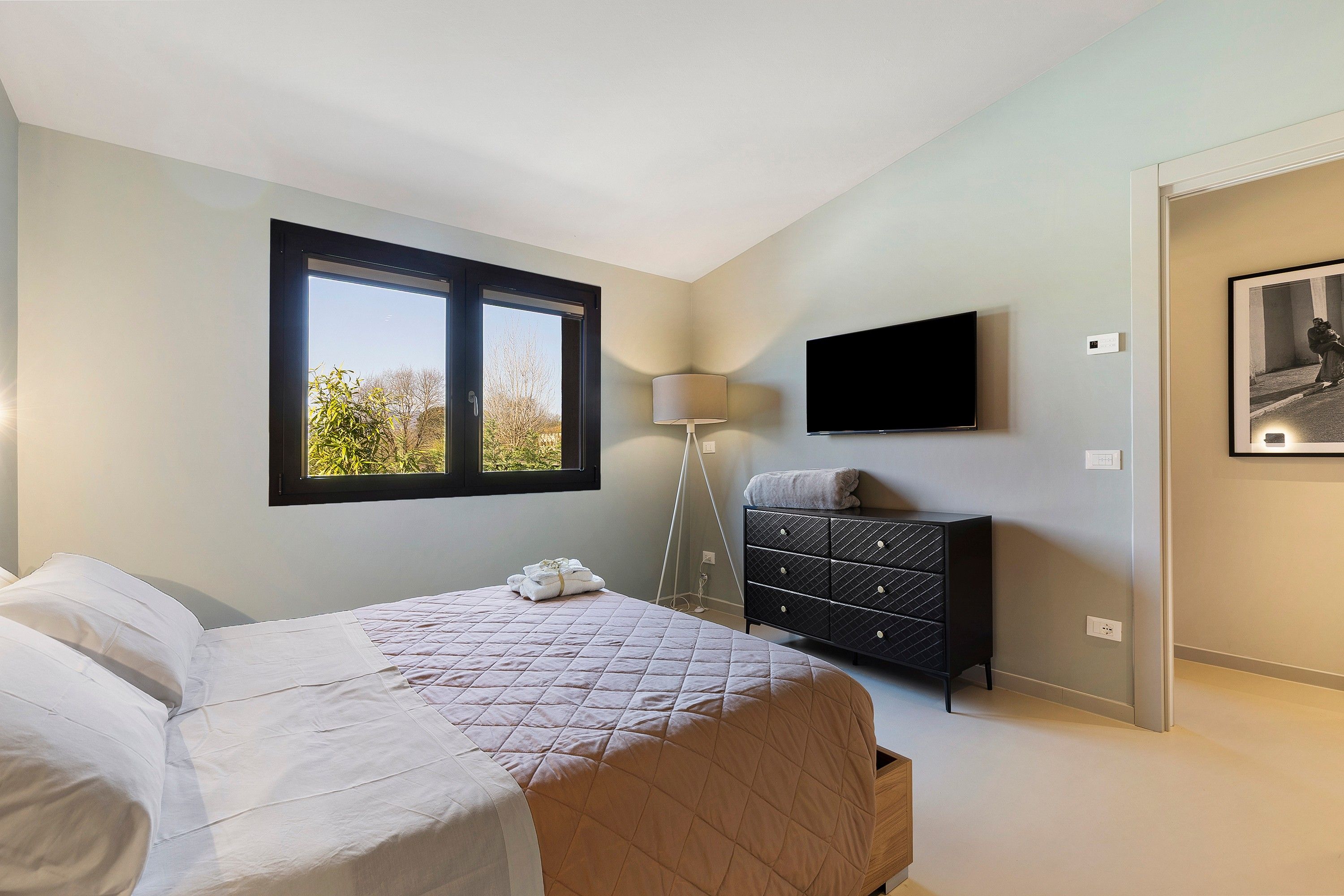 Ein Schlafzimmer mit Bett, Kommode, Lampe, Fernseher und Fenster.