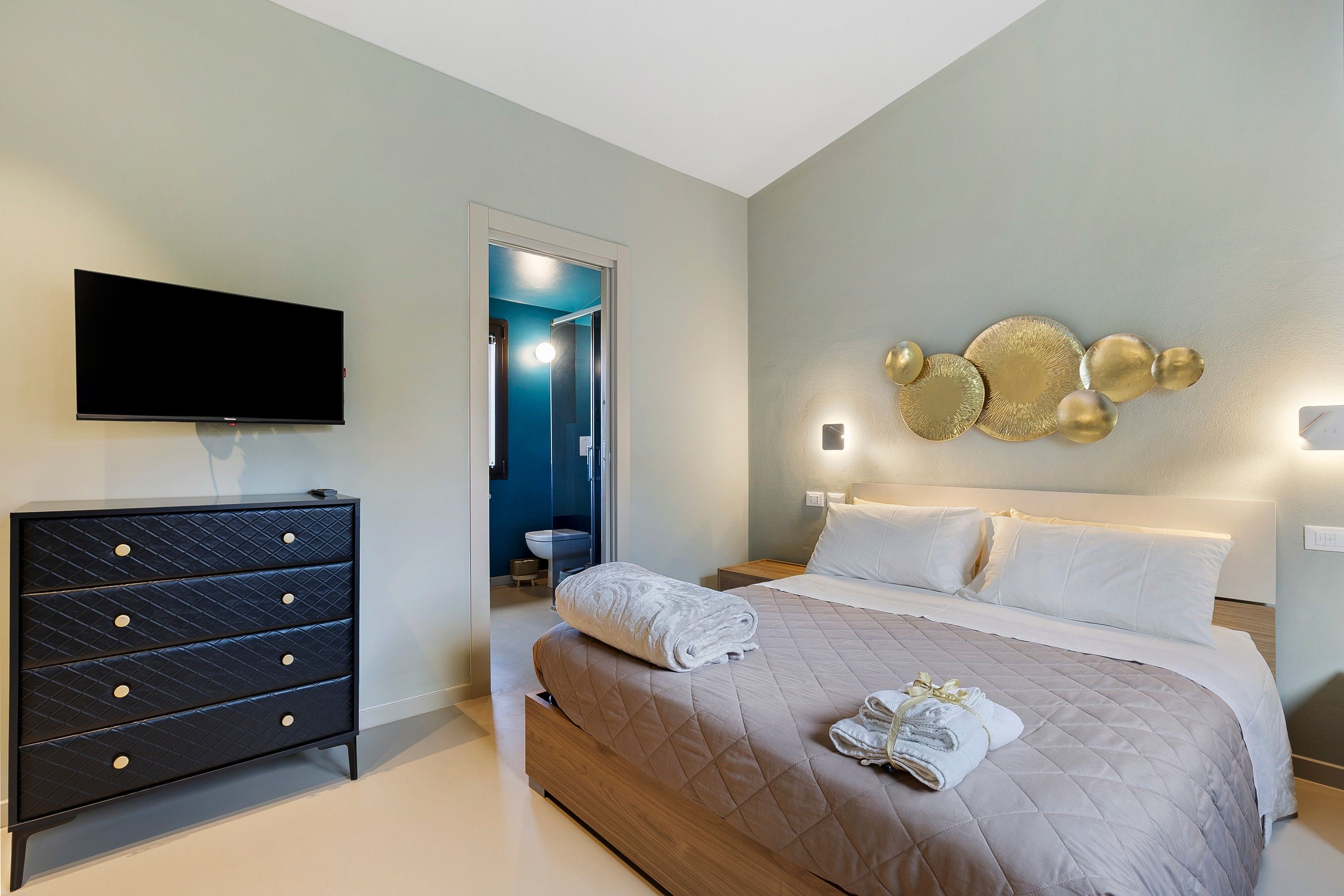 A bedroom with a bed, a dresser, a flat-screen TV, and a wall-mounted light fixture.