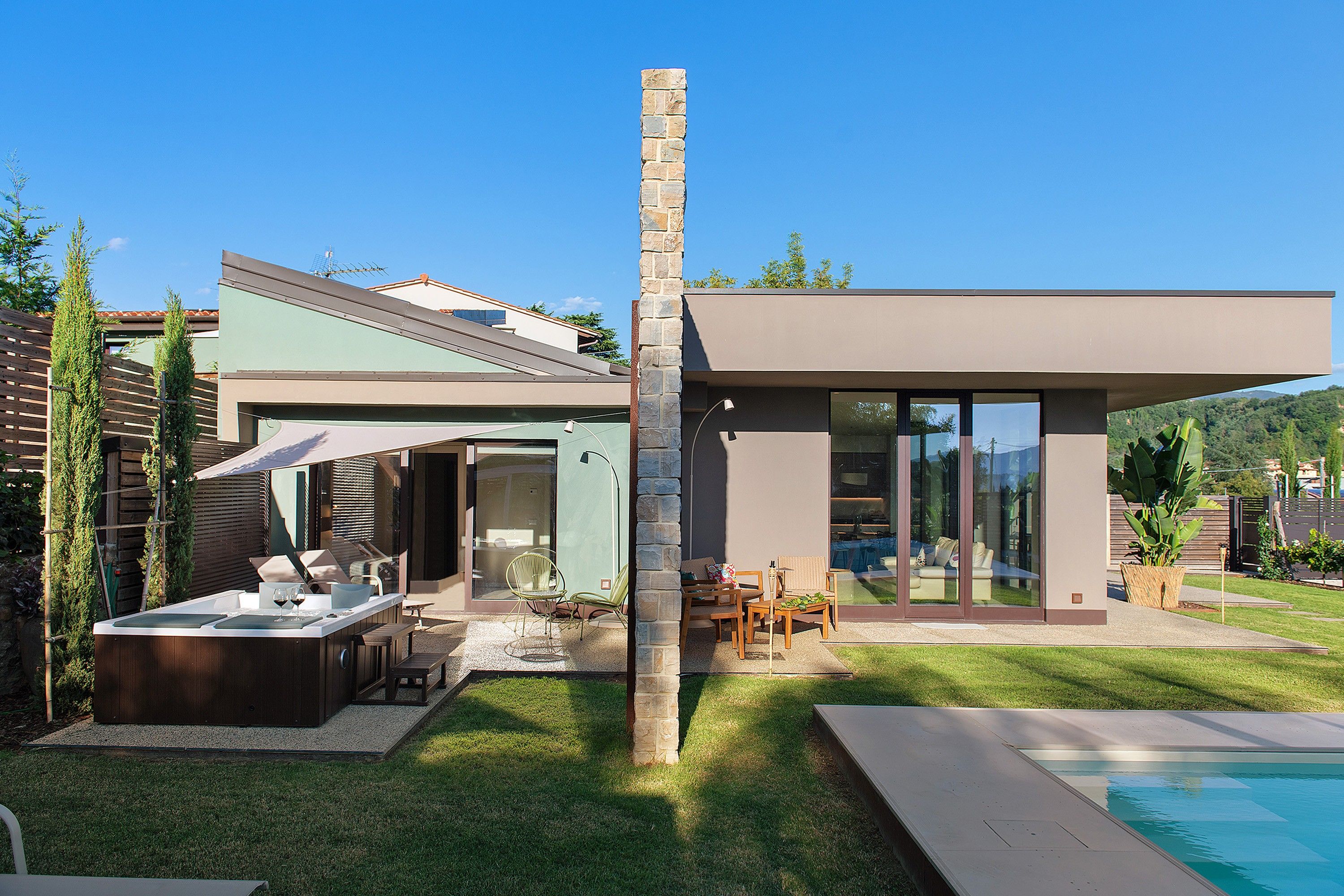 A modern house with a stone pillar, a swimming pool, and outdoor furniture.