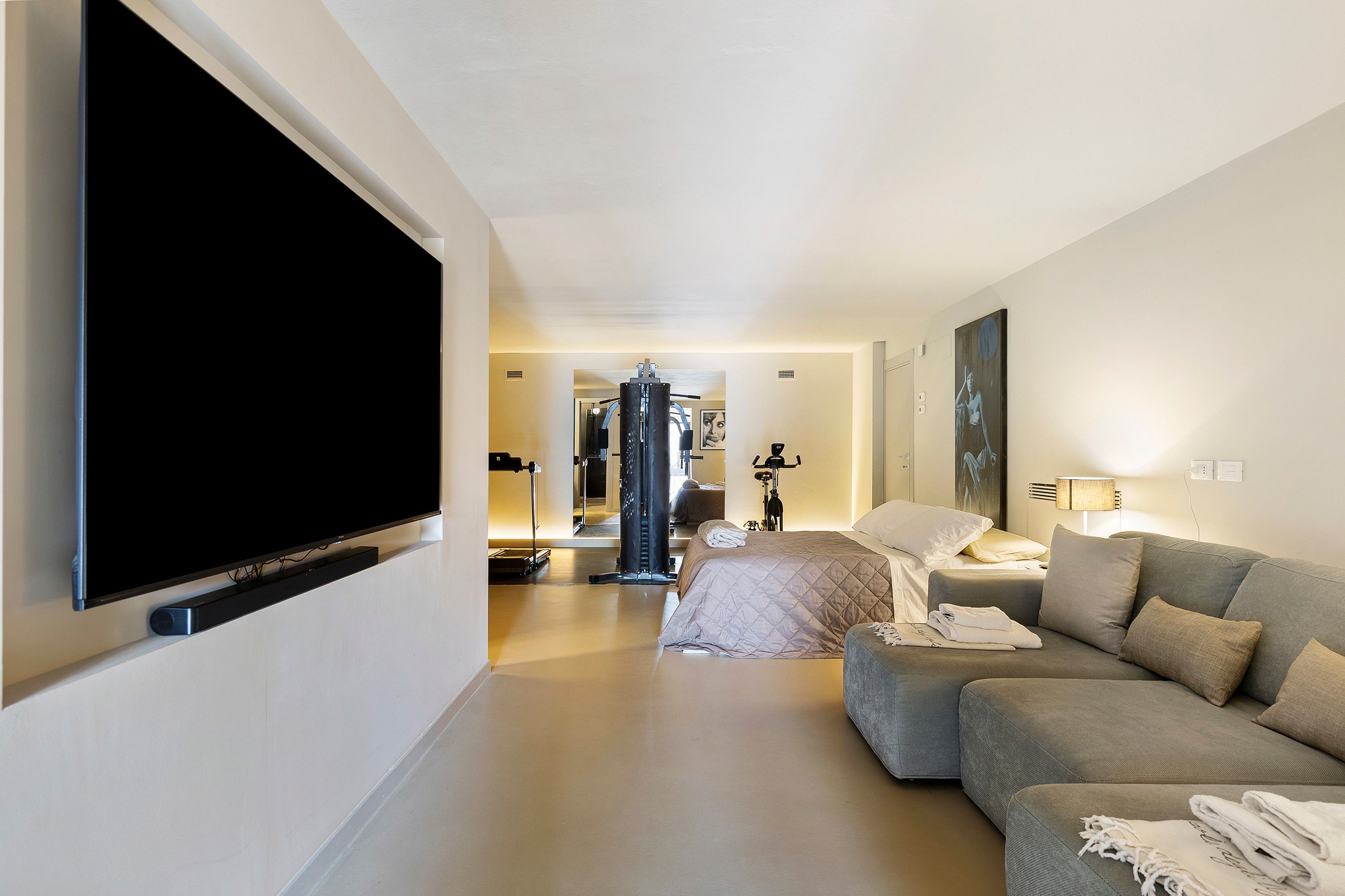 Ein modernes Hotelzimmer mit Flachbildfernseher, Sofa, Bett und Trainingsgerät, sowie einem Gemälde an der Wand.