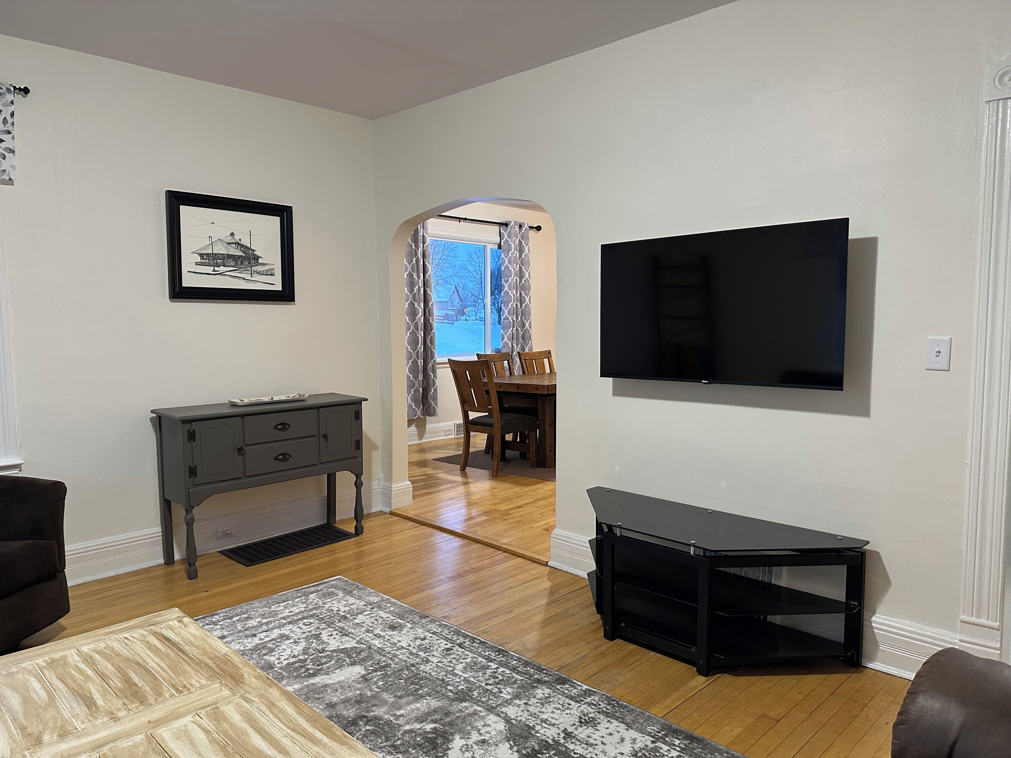 A living room with a flat-screen TV, a gray console table, a patterned rug, and a dining area with a wooden table and chairs.