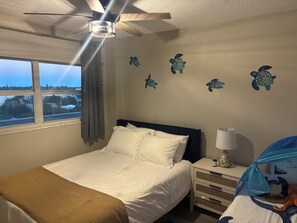 Ein Hotelzimmer mit Bett, Nachttisch, Lampe, einem Fenster mit Vorhängen und einem Deckenventilator.