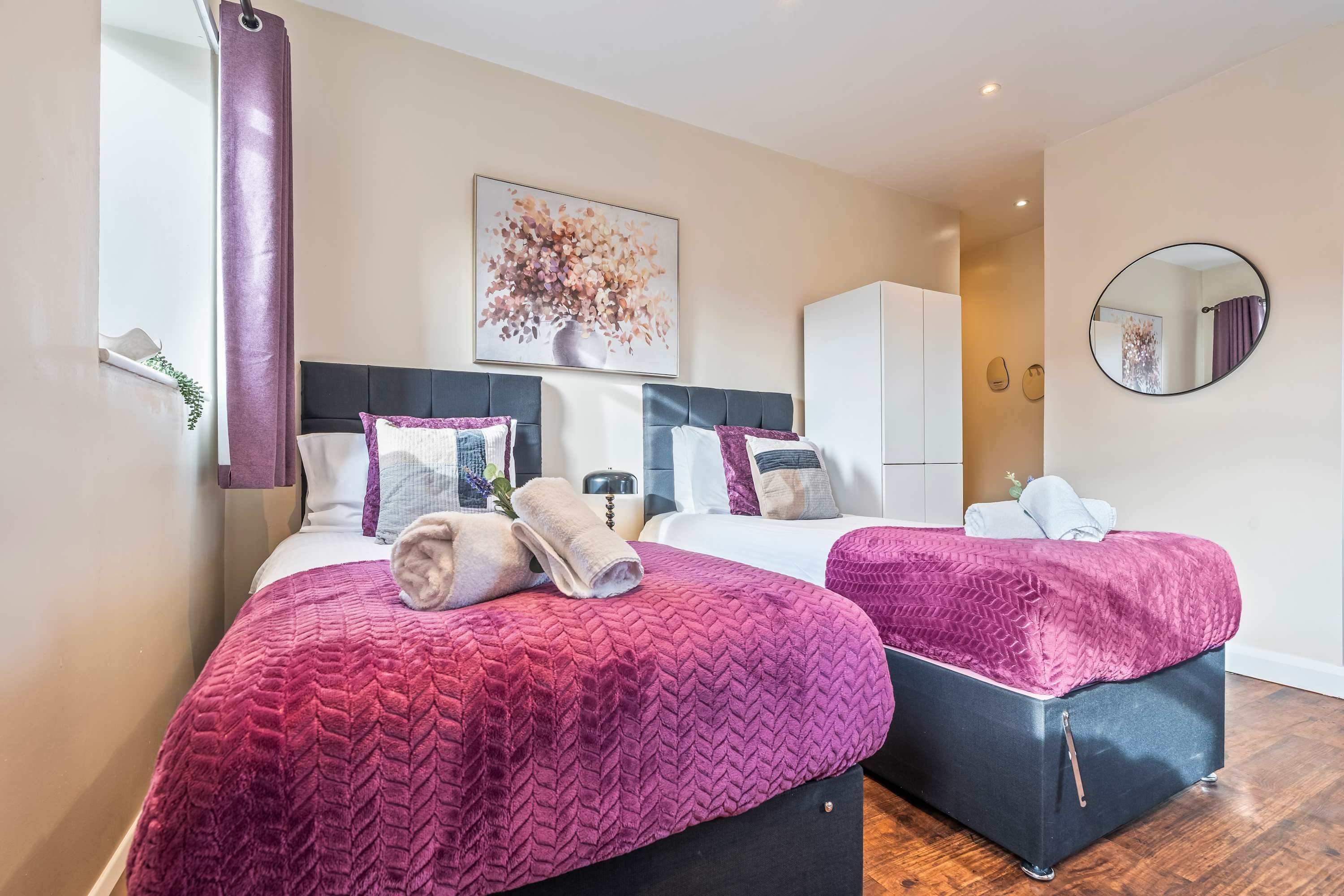 Habitación de hotel con dos camas, un ventanal con cortinas, un cuadro en la pared y un espejo redondo.