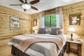 Ein Schlafzimmer mit Holzwandverkleidung, einem Bett, zwei Nachttischen, einem Deckenventilator und einem Fenster mit Vorhängen.