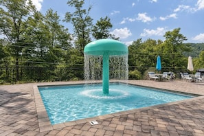A swimming pool with a water fountain feature, surrounded by a brick deck and lounge chairs. - Smoky Mtn Escape Hot Tub Fire Pit Game Room (Gatlinburg)