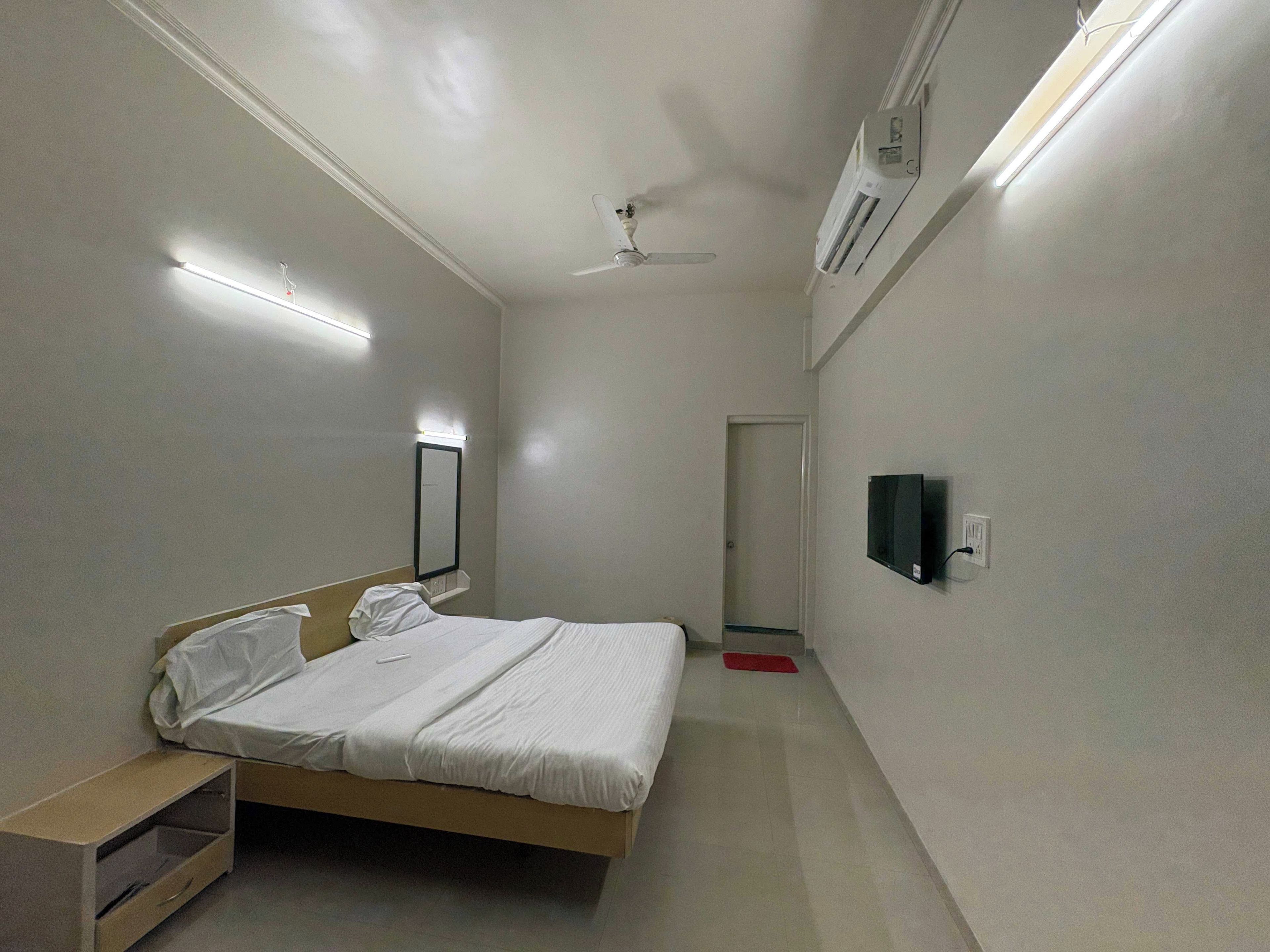 Un dormitorio pequeño y bien iluminado con una cama individual, una mesita de noche, un espejo, un ventilador de techo y un televisor montado.