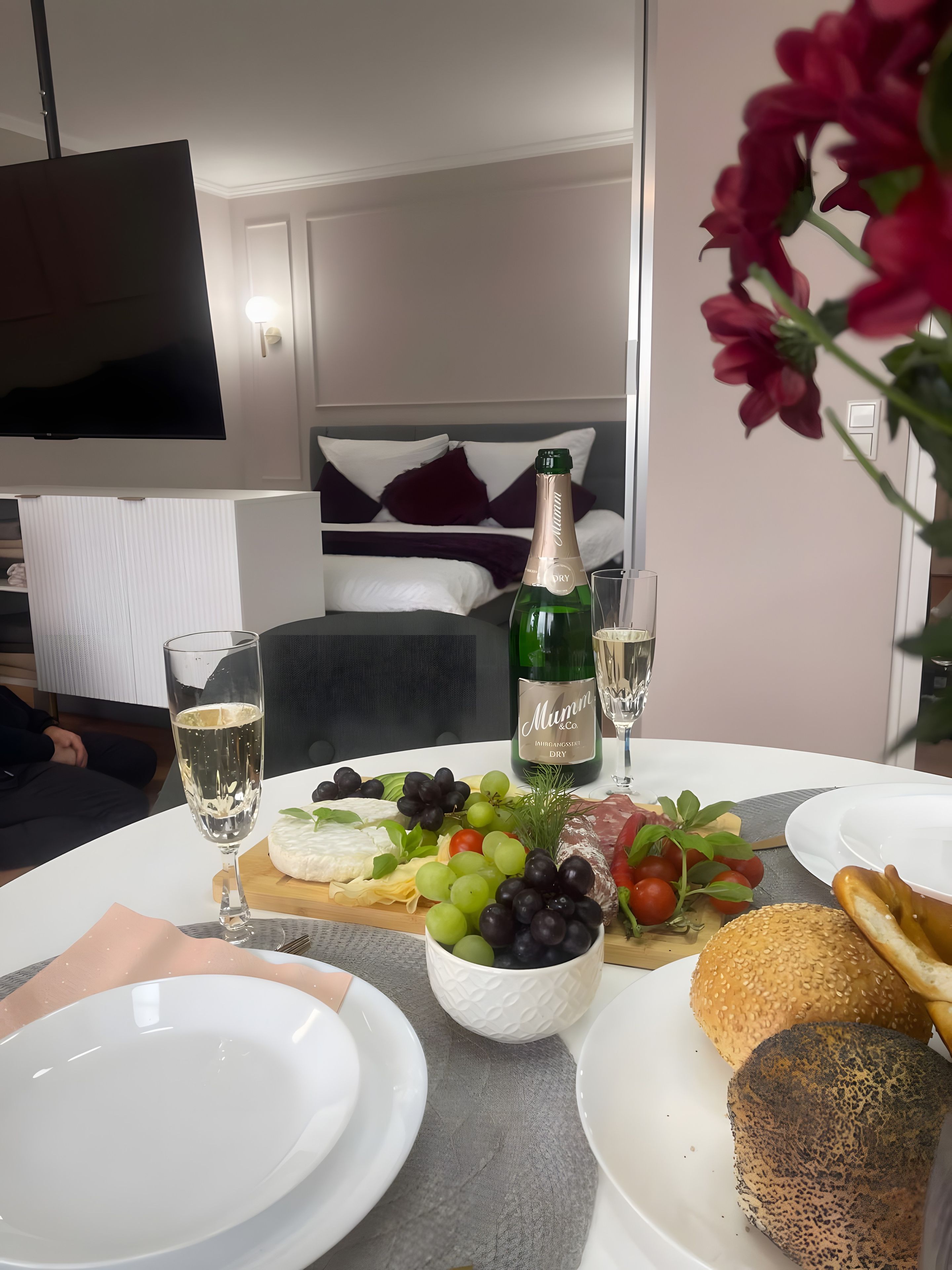 A dining table with a bottle of champagne, glasses, a cheese board, and various fruits.