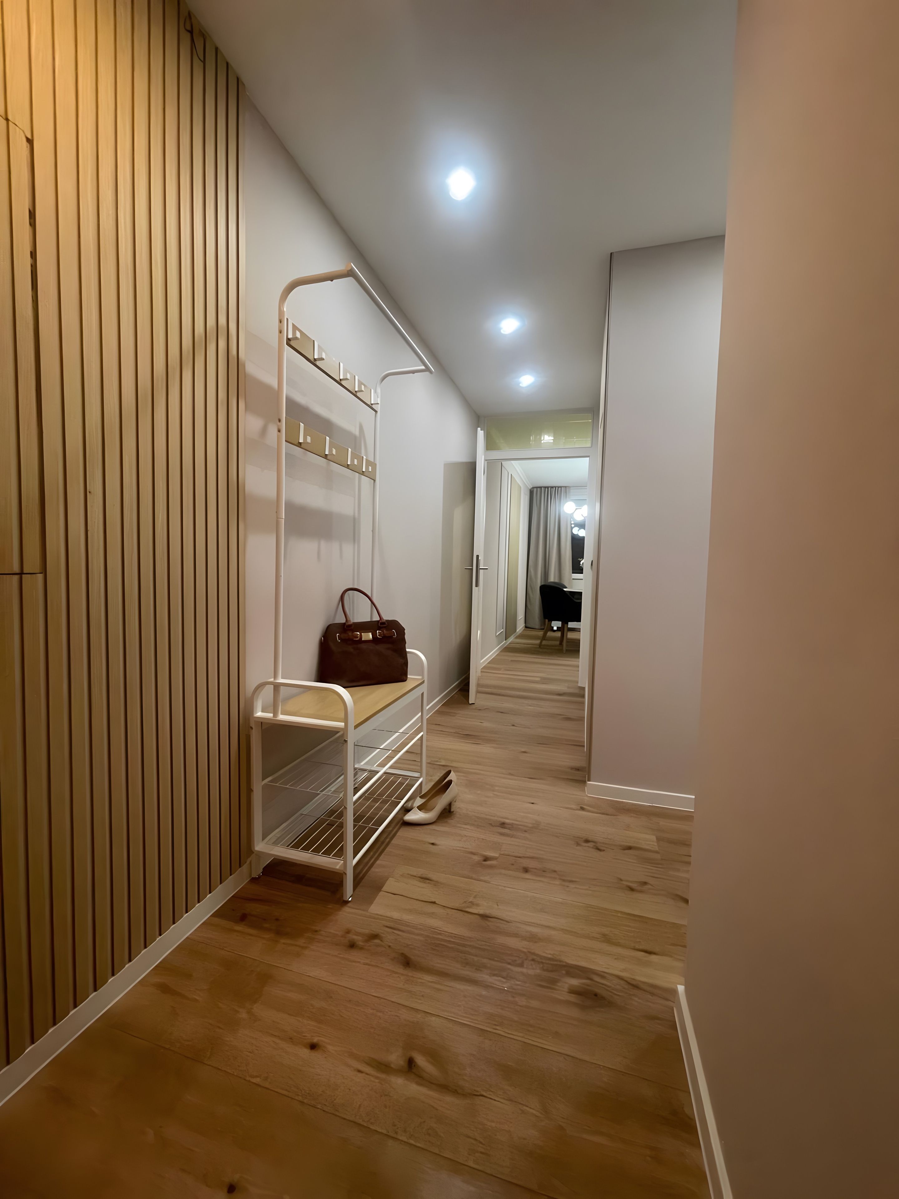 A hallway with a wooden floor, a white metal bench with a handbag, and a door leading to another room.
