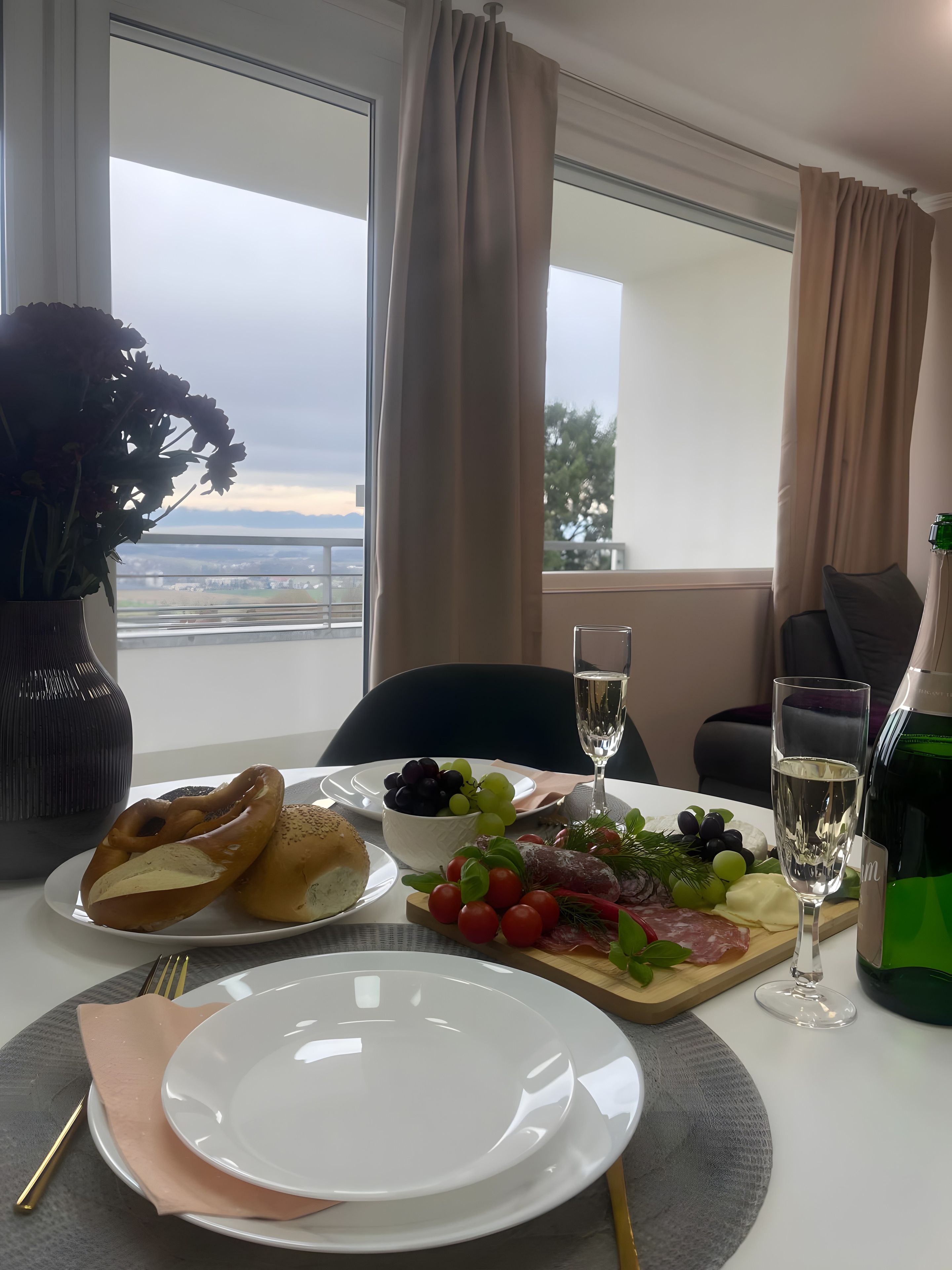 Une table dressée avec des bretzels, des raisins, du fromage et une bouteille de vin, avec une vue sur la ville en arrière-plan.