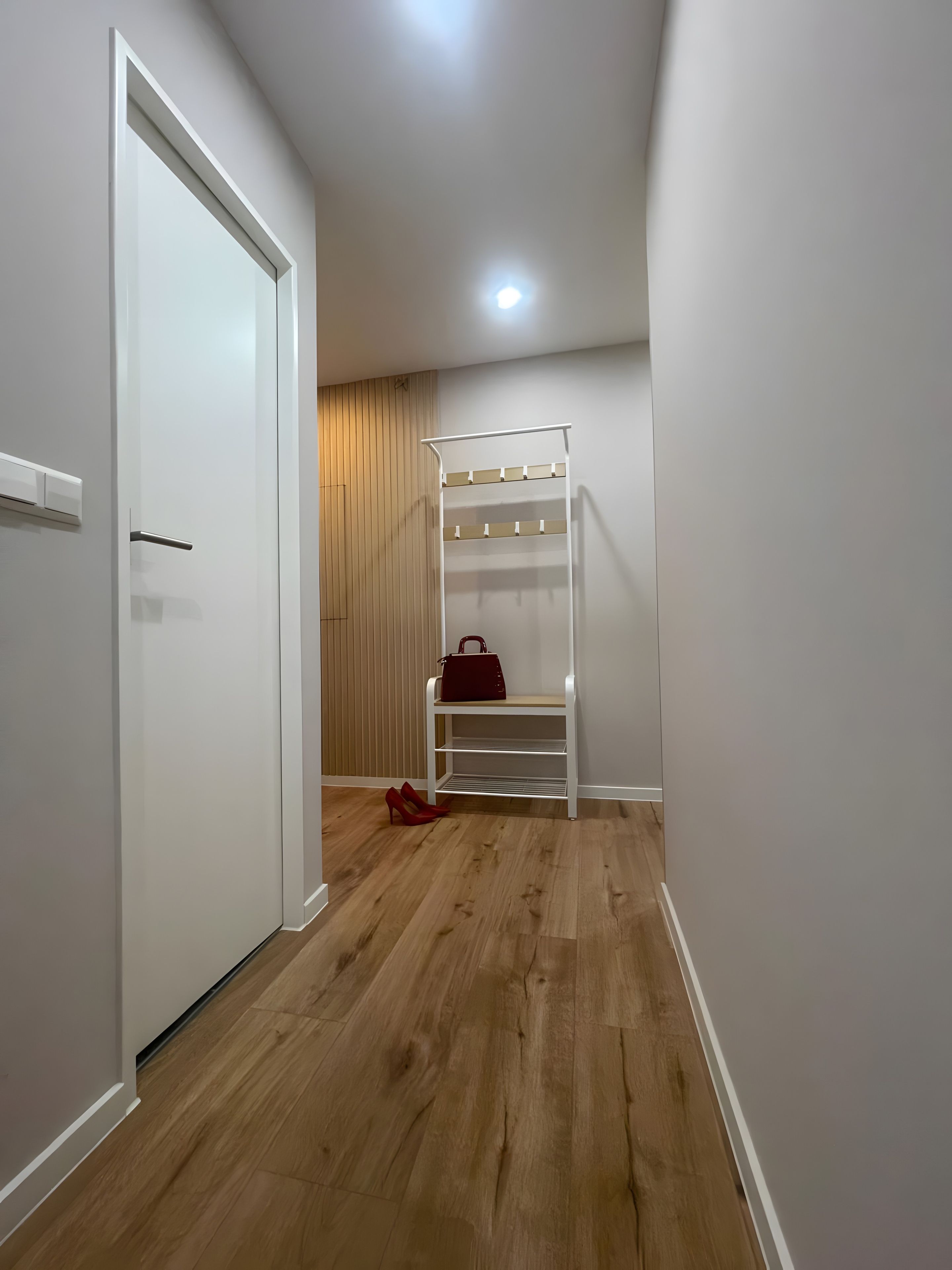 Un couloir avec une porte blanche, un plancher en bois et une étagère blanche sur laquelle se trouvent un sac à main et une paire de chaussures rouges.