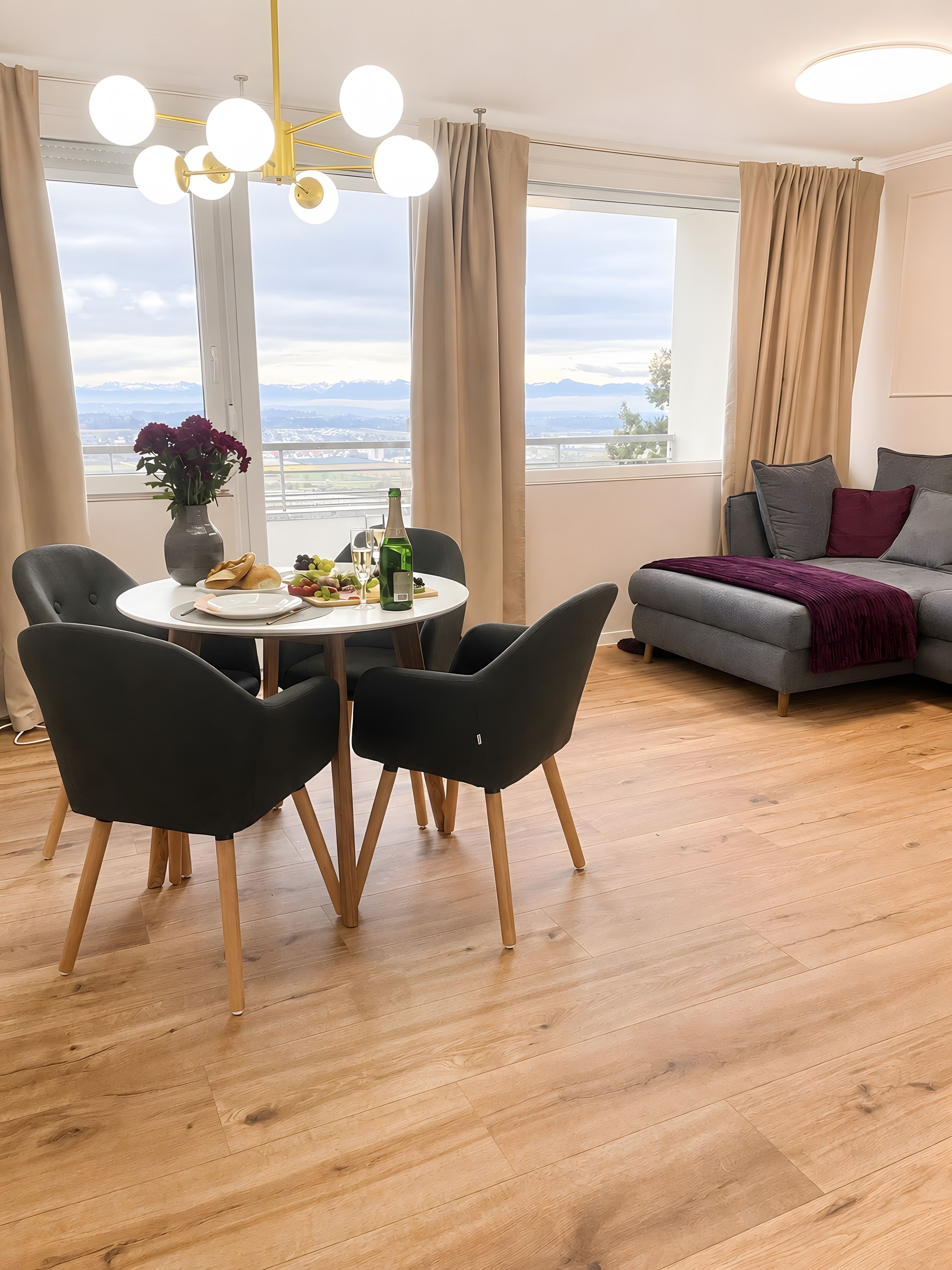 Sala de estar moderna com área de jantar, janelas amplas com cortinas, sofá cinza e lustre.