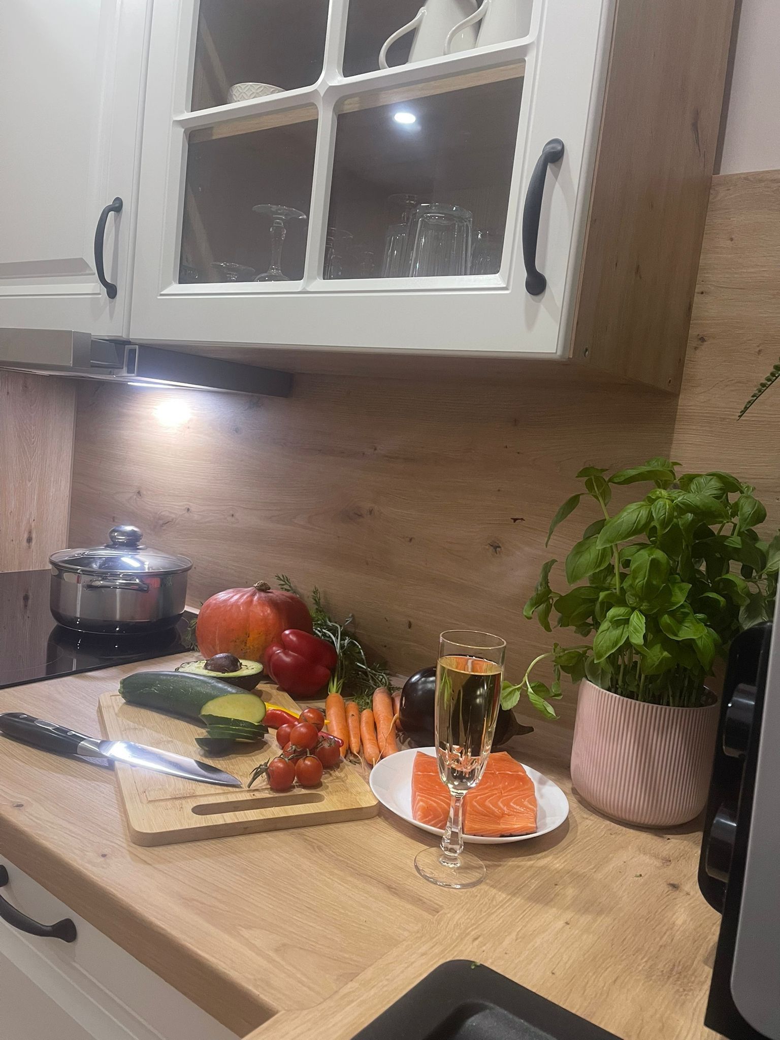 Eine Küchenarbeitsplatte mit Schneidebrett, Gemüse, einem Glas Wasser, einem Teller mit Lachs und einer Topfpflanze.