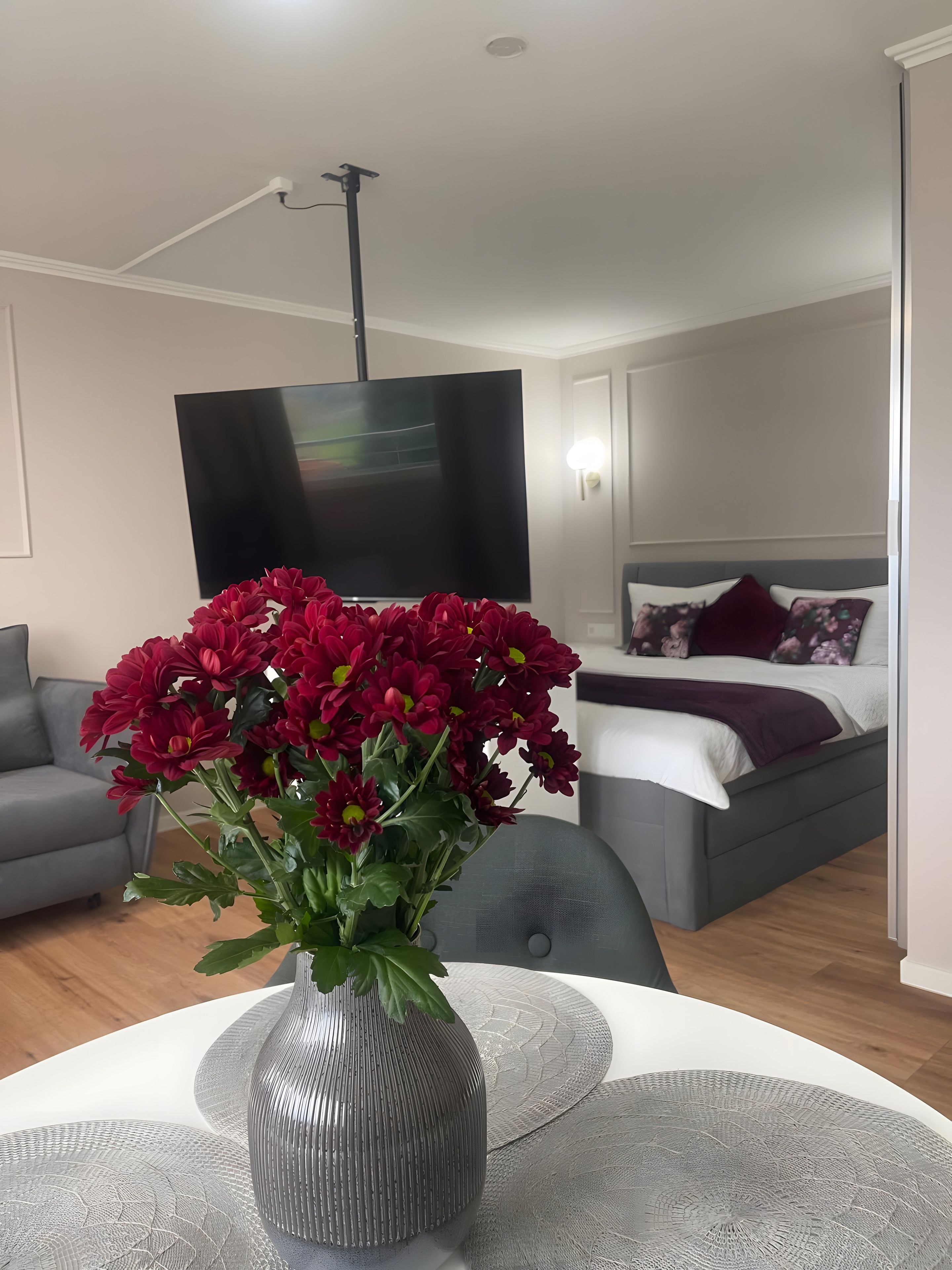 A modern living room with a grey sofa, a bed with a grey headboard, a flat-screen TV mounted on the wall, and a vase with red flowers on a white table.
