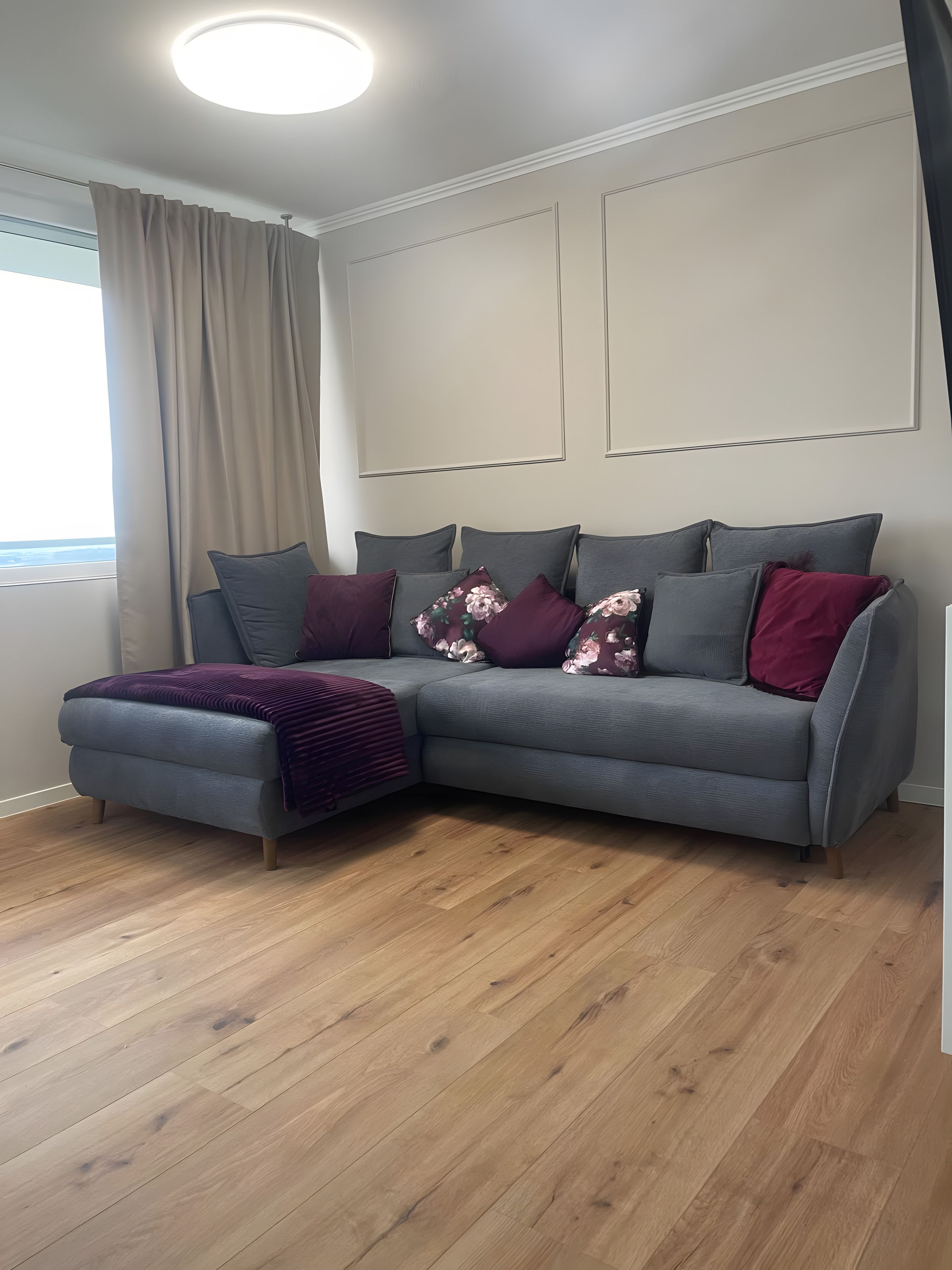 Ein modernes Wohnzimmer mit einem grauen Sofa, Holzboden und einem Fenster mit Vorhängen.