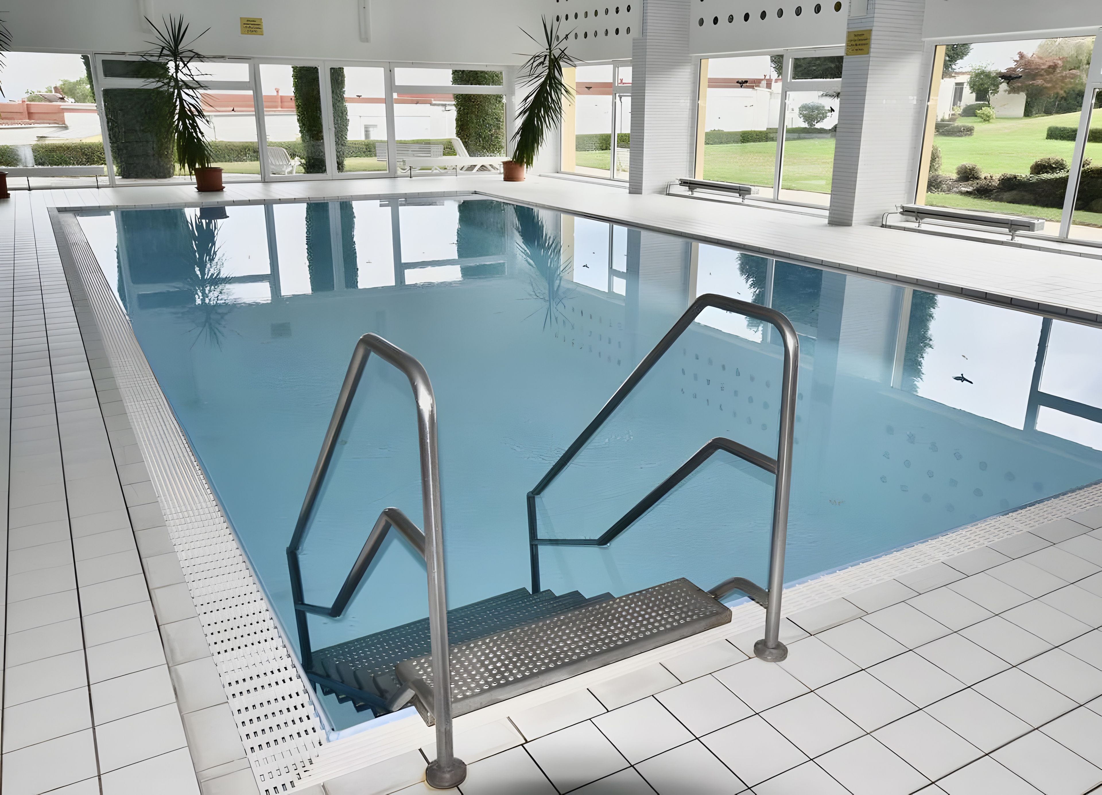 Une piscine intérieure avec des parois en verre transparent, des échelles métalliques et des plantes en pot.
