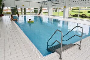 An indoor swimming pool with clear blue water, a tiled floor, and a view of the outdoors through large windows. - Apartment 'See und Bergblick 58 mit Indoorpool' (Oberteuringen)