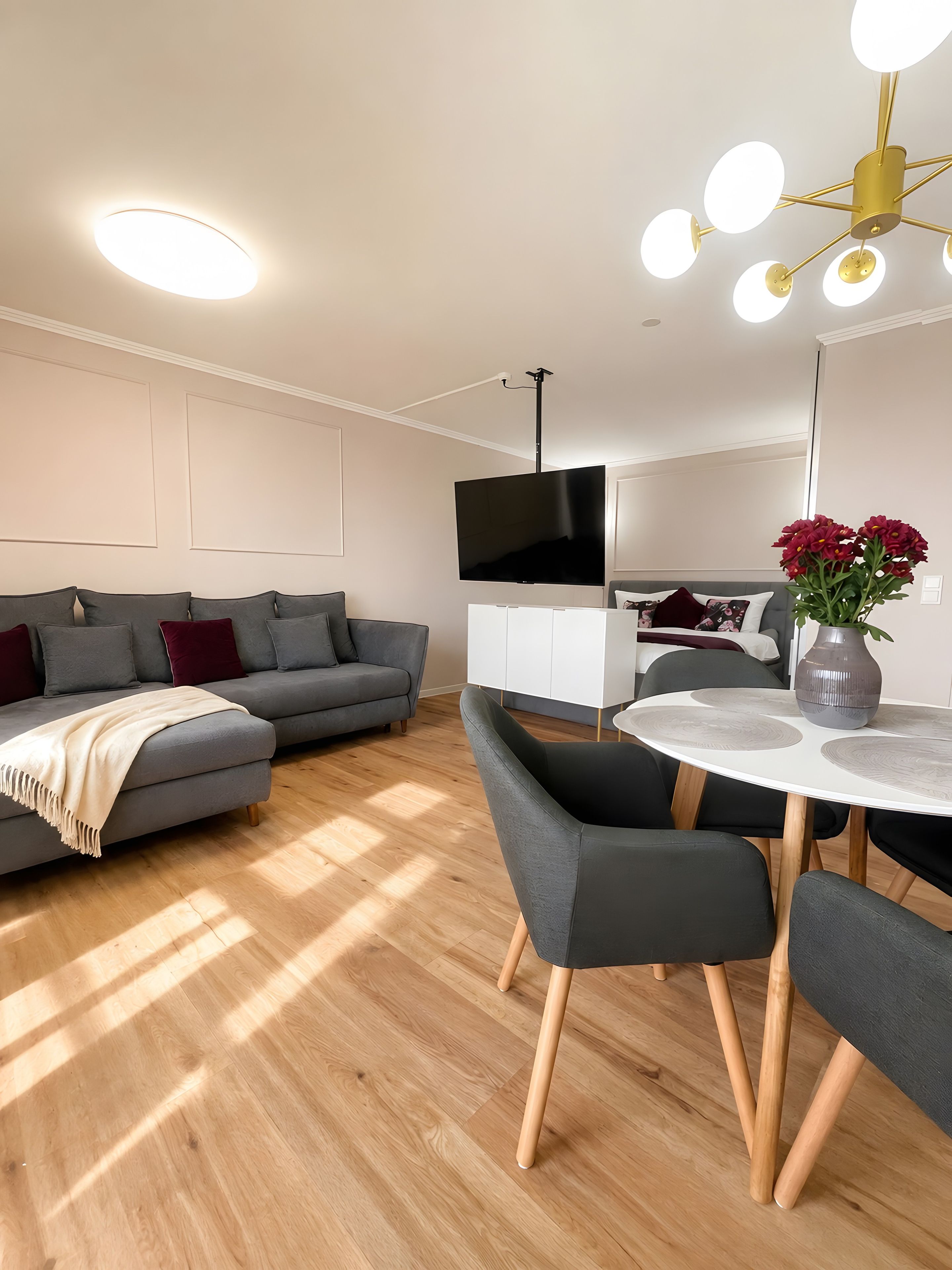 A modern living room with a grey sofa, a flat-screen TV, a dining table with chairs, and a ceiling light fixture.