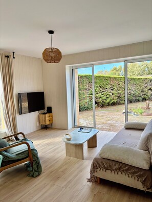 A modern living room with a sofa, a coffee table, a TV, and a large window offering a view of a garden. - An Autumn Getaway in Cap Ferret, Amid Pine Trees and the Lagoon (Lège-Cap-Ferret)