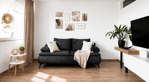 Una sala moderna con un sofá oscuro, una mesita auxiliar, una planta en maceta y un televisor de pantalla plana sobre un soporte blanco.