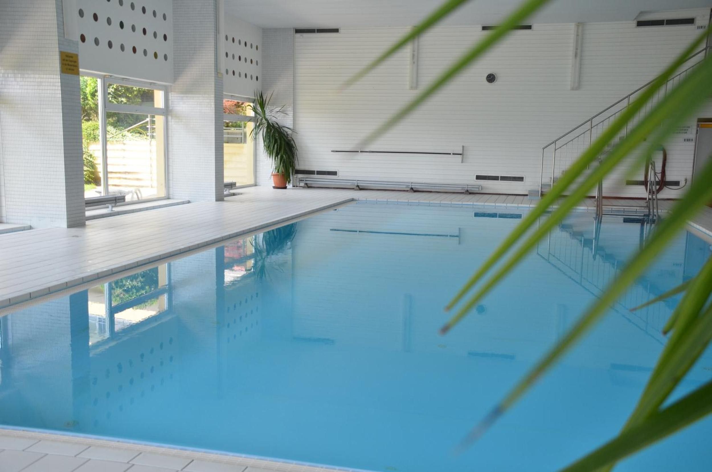 Ein beheiztes Hallenbecken mit klarem, blauem Wasser, umgeben von weißen Wänden und einem gefliesten Boden.