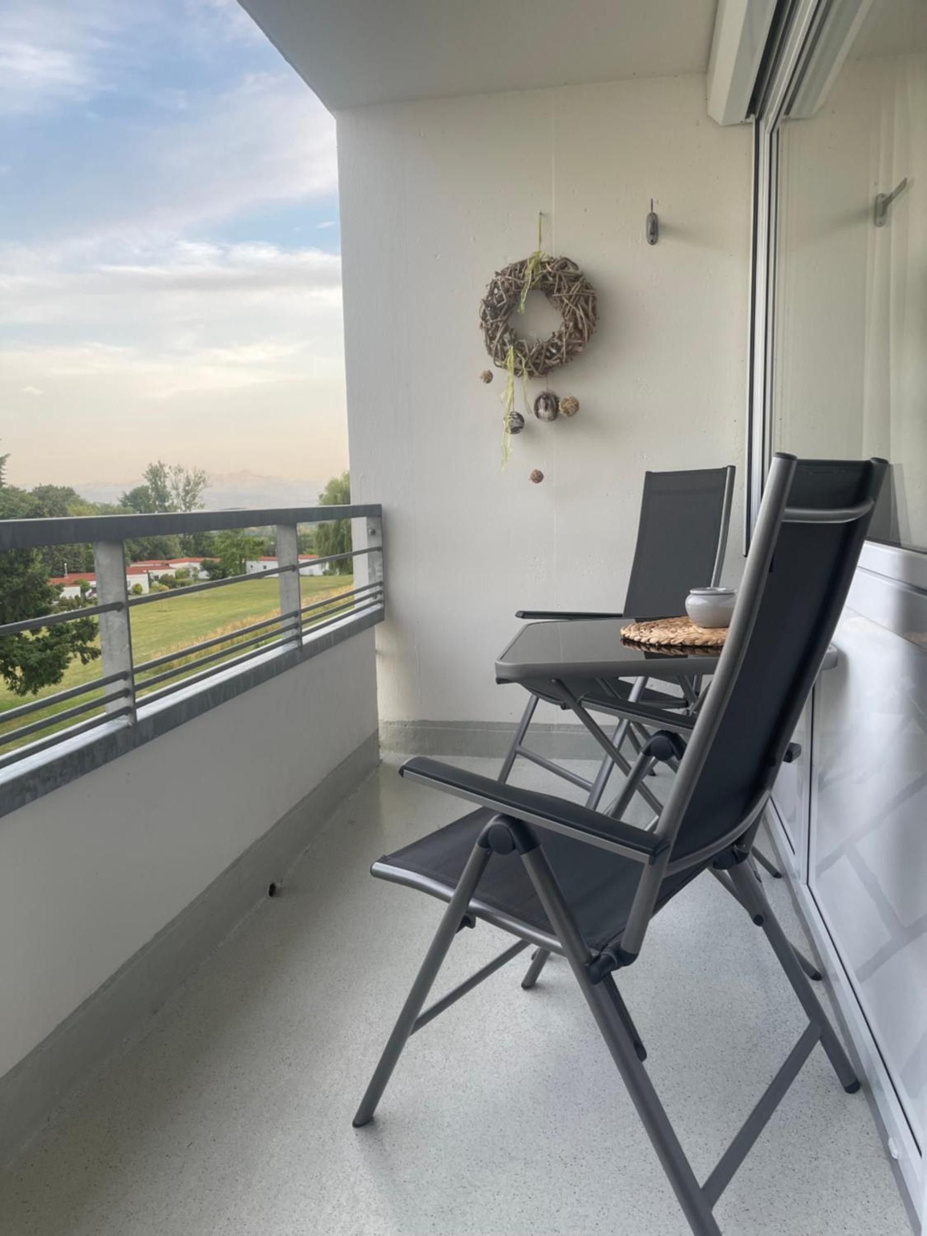 A balcony with a view of a green landscape, a metal railing, a table with a chair, and a decorative wreath on the wall.