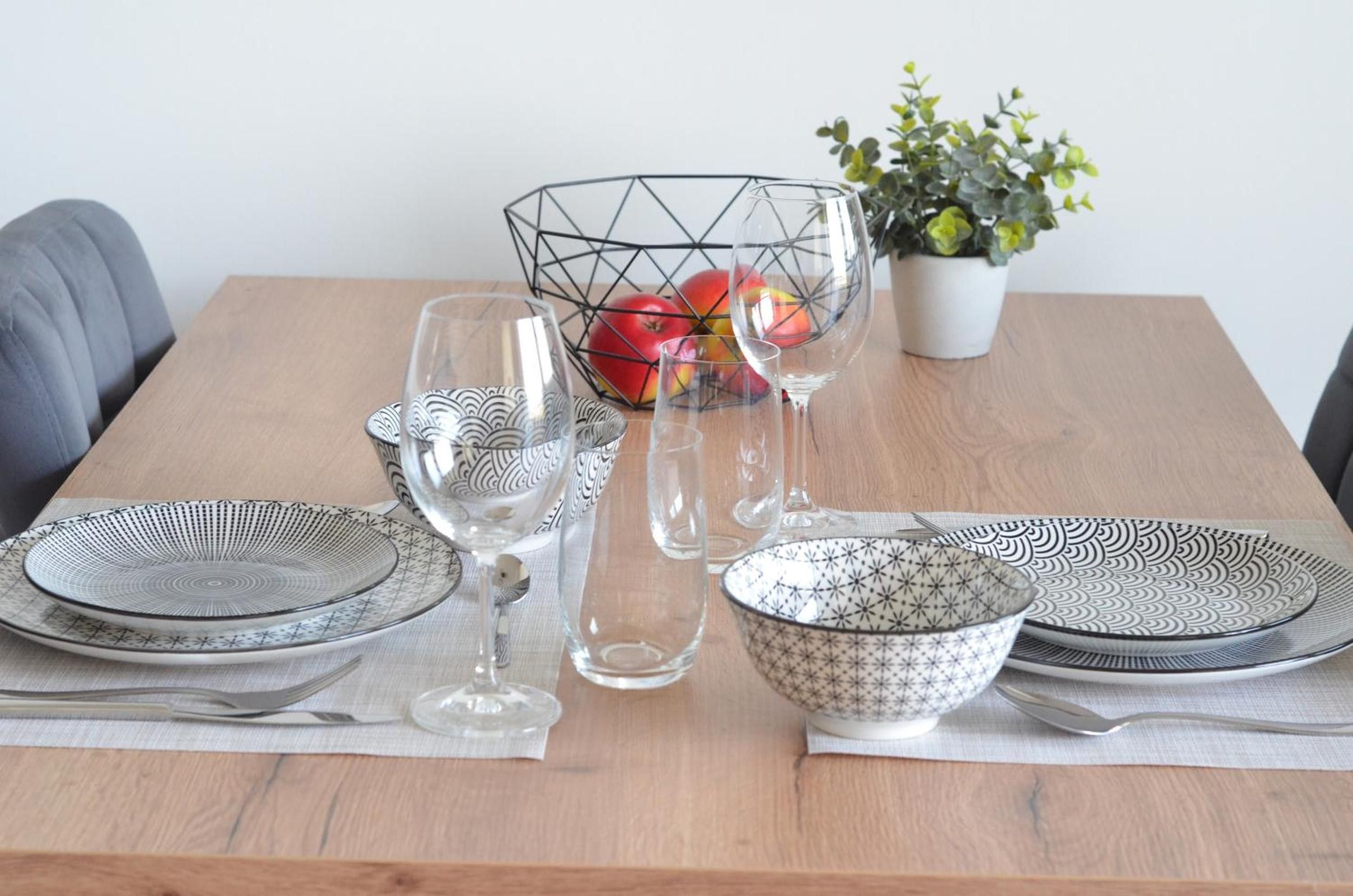 A dining table set with plates, bowls, and glasses, with apples in a wire basket and a potted plant in the background.