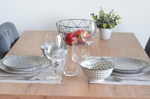 A dining table set with plates, bowls, and glasses, with apples in a wire basket and a potted plant in the background.