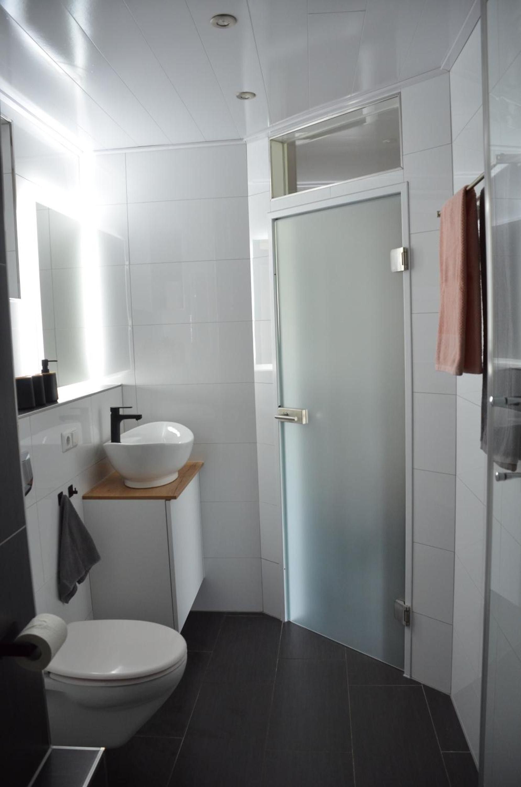 Une salle de bain moderne avec des toilettes blanches, un lavabo blanc posé sur une étagère en bois, une cabine de douche en verre et une serviette accrochée à la porte.