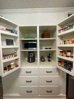 Una cocina con gabinetes blancos, microondas, cafetera y varios condimentos y especias en estantes.
