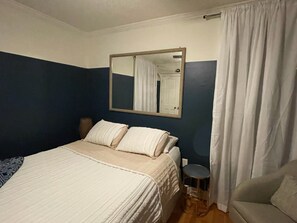 Un dormitorio con una cama, una mesita de noche, una silla, un espejo y una cortina.