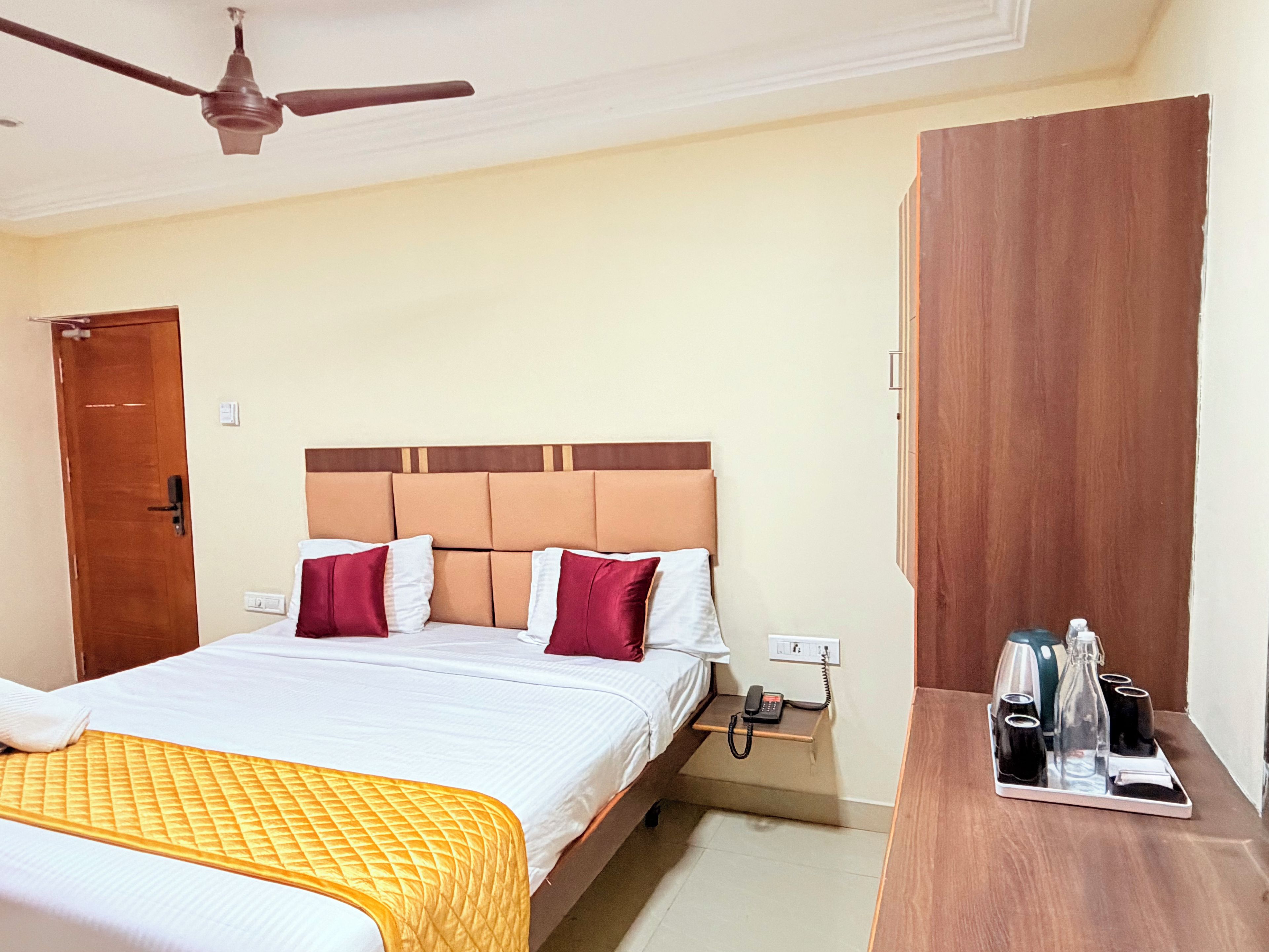 A hotel room with a bed, a bedside table with a phone and a tray with glasses and a bottle, a ceiling fan, and a wooden wardrobe.
