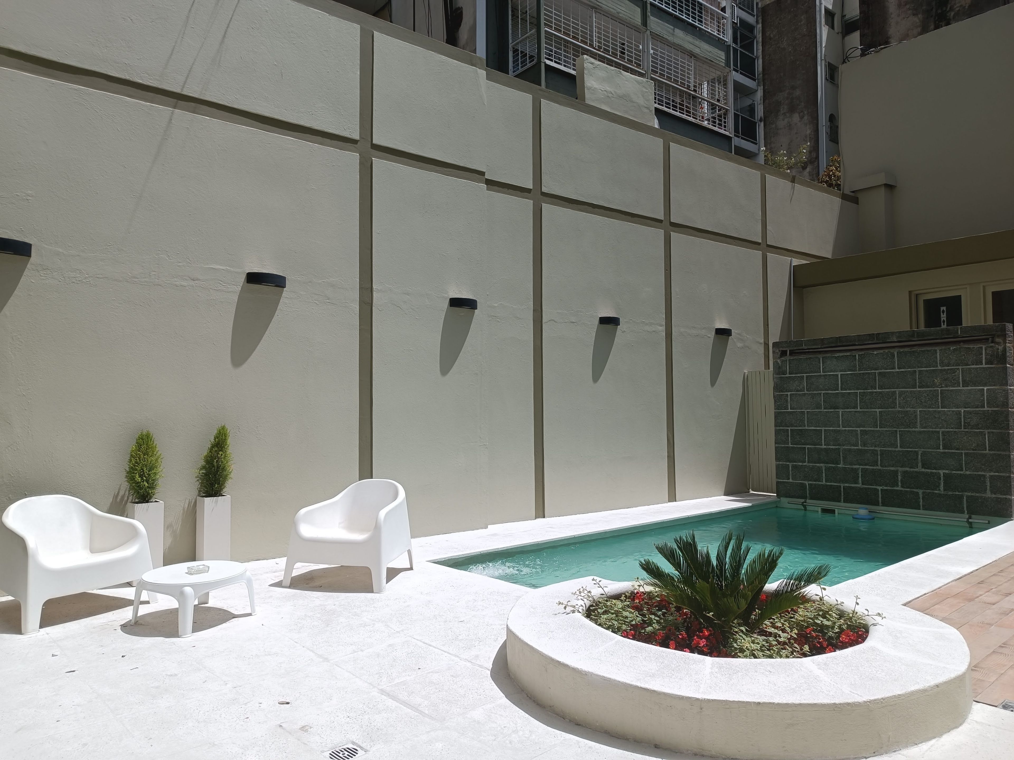 Un espace aménagé au bord de la piscine, avec une piscine blanche, des chaises blanches, une petite table et un bac à fleurs orné de fleurs rouges.
